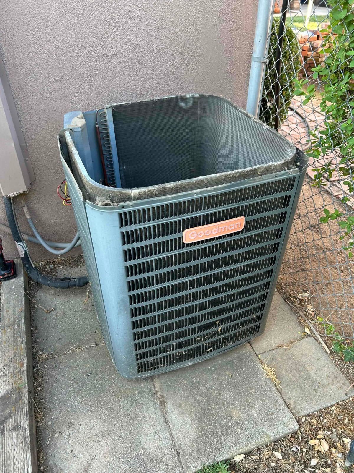 Old outdoor air conditioner unit beside a house wall and fence, with a bent top grill.