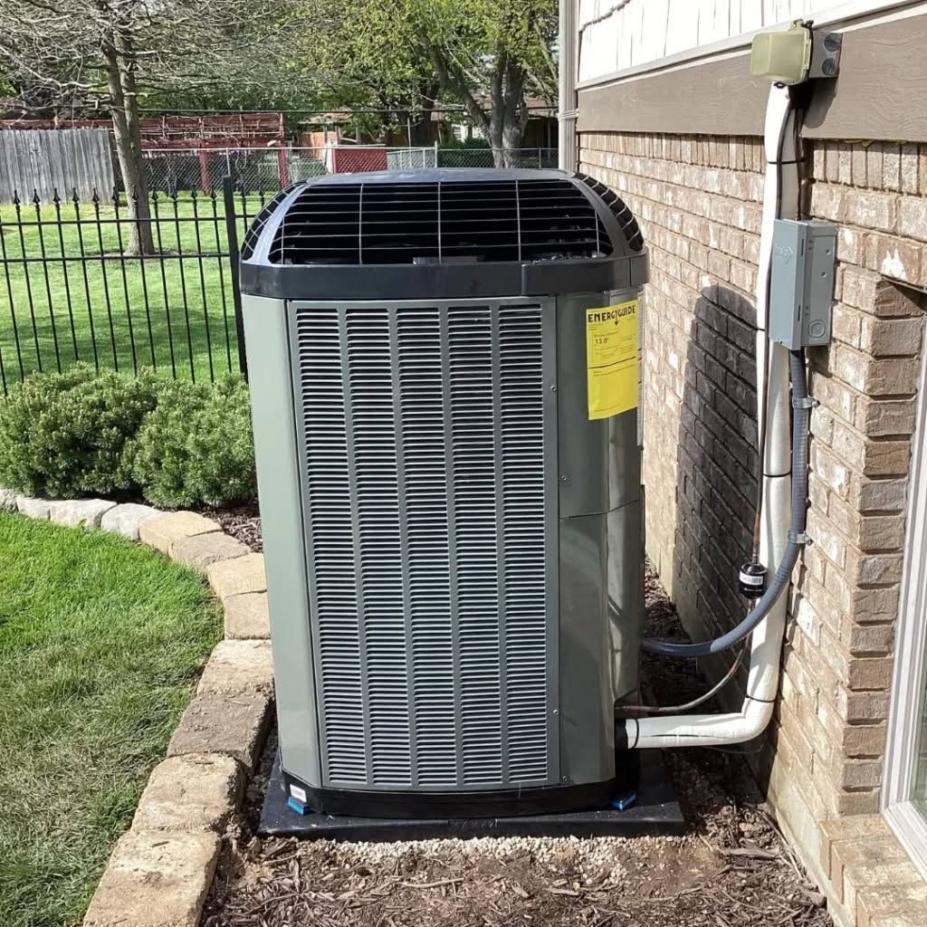Outdoor air conditioner unit beside a brick house, with a fence and lawn in the background.