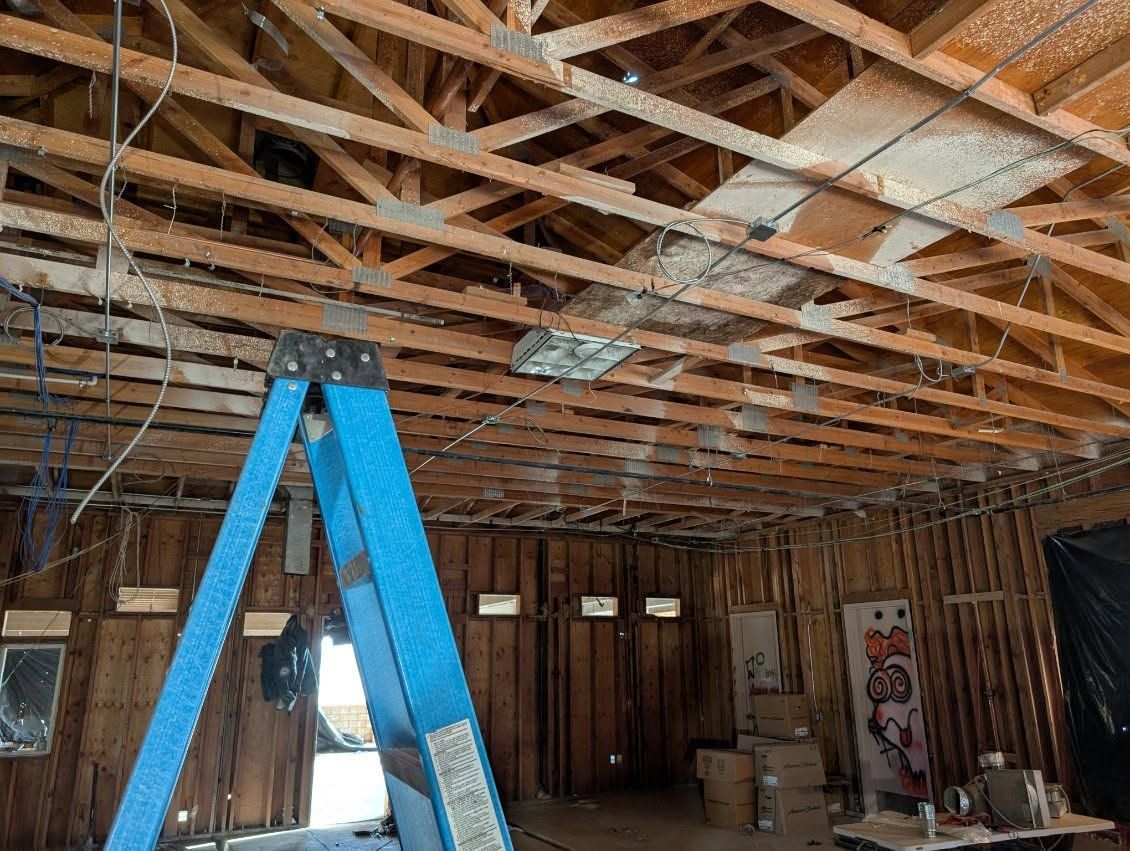 Construction site with exposed wooden ceiling joists and a blue ladder in the foreground.