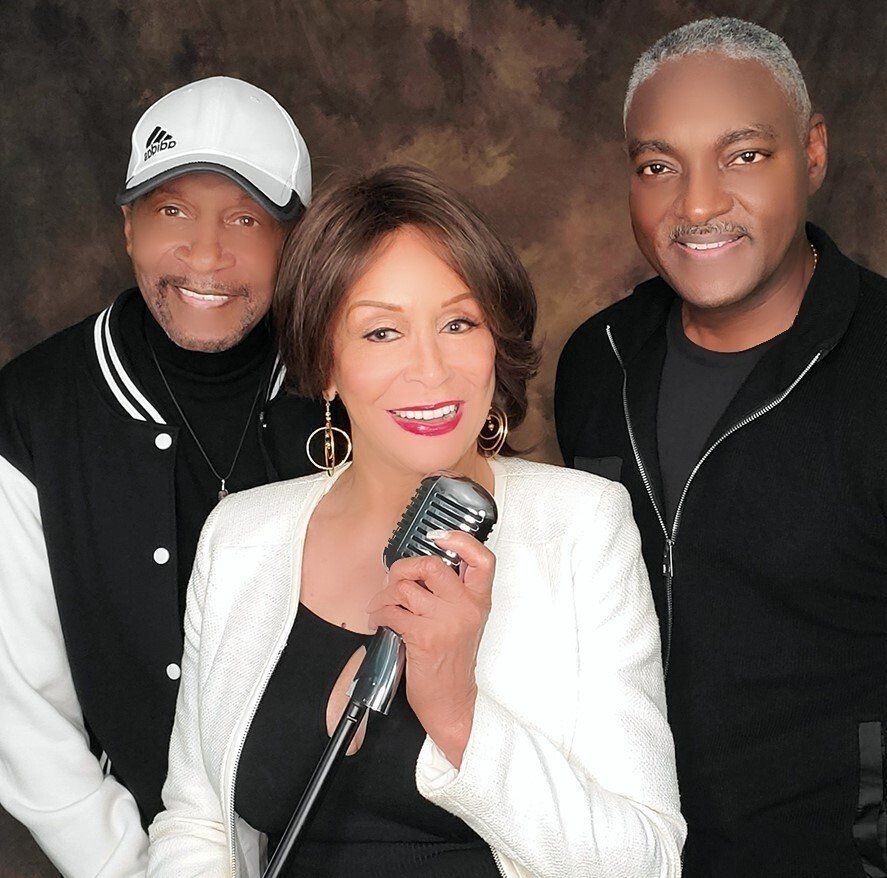 Three singers posing with microphones, one in front wearing a white jacket and smiling.