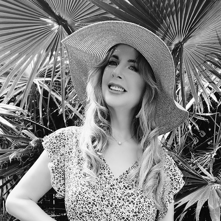 A woman wearing a straw hat and a floral dress is standing in front of a palm tree.