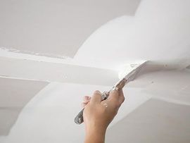 Hand using a putty knife to smooth white joint compound on a ceiling seam