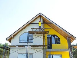 Worker on scaffolding painting a white house with yellow trim under a steep roof