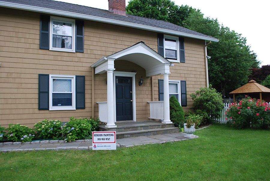 Two-story tan house with a front porch, steps, lawn, and landscaping