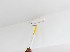 Hand holding a paint roller applying white paint to a ceiling corner