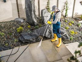Person pressure washing a concrete patio beside rocks and trees, wearing yellow boots and gloves.