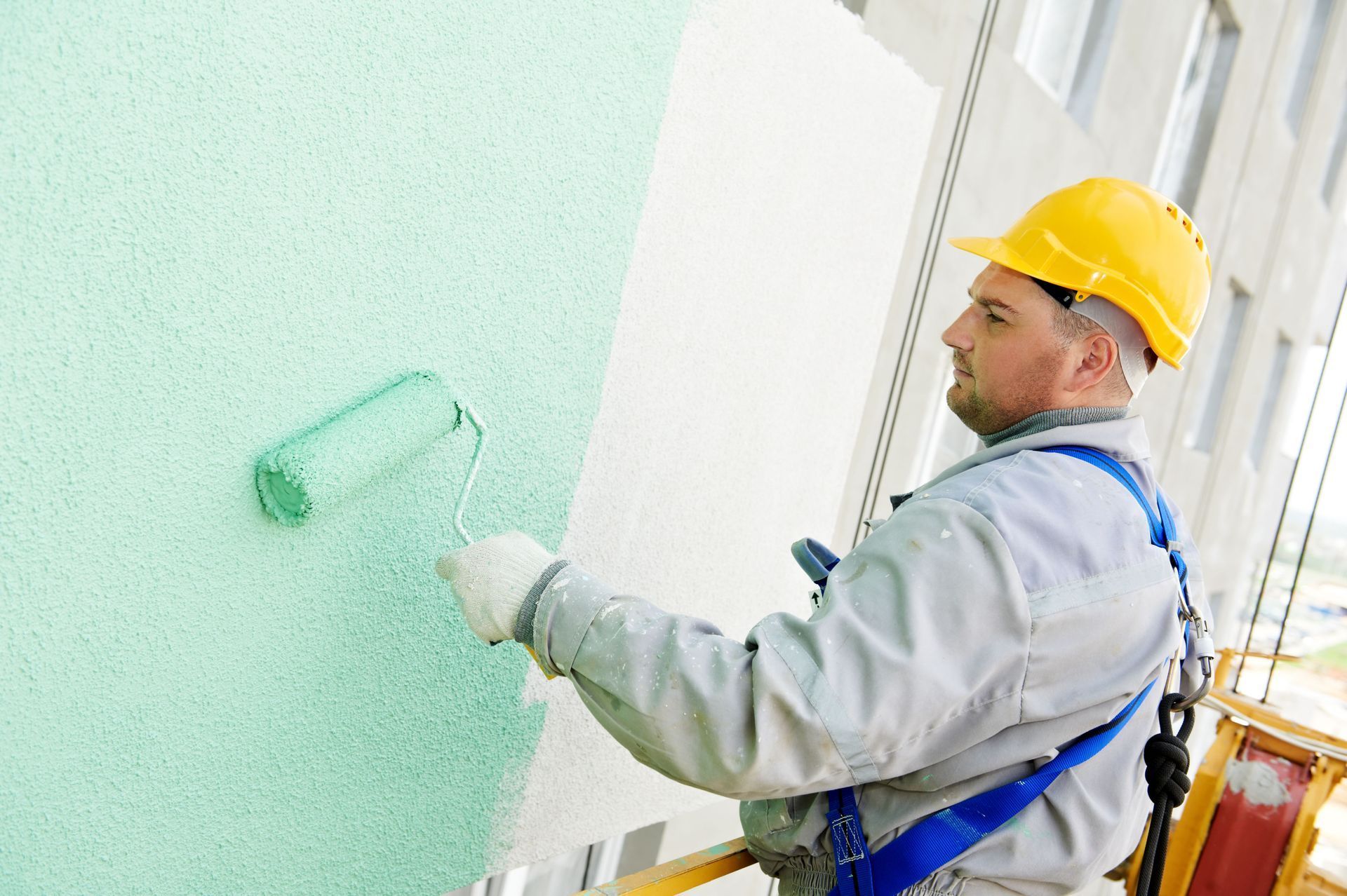 Worker in a yellow hard hat rolling light green paint onto a wall.