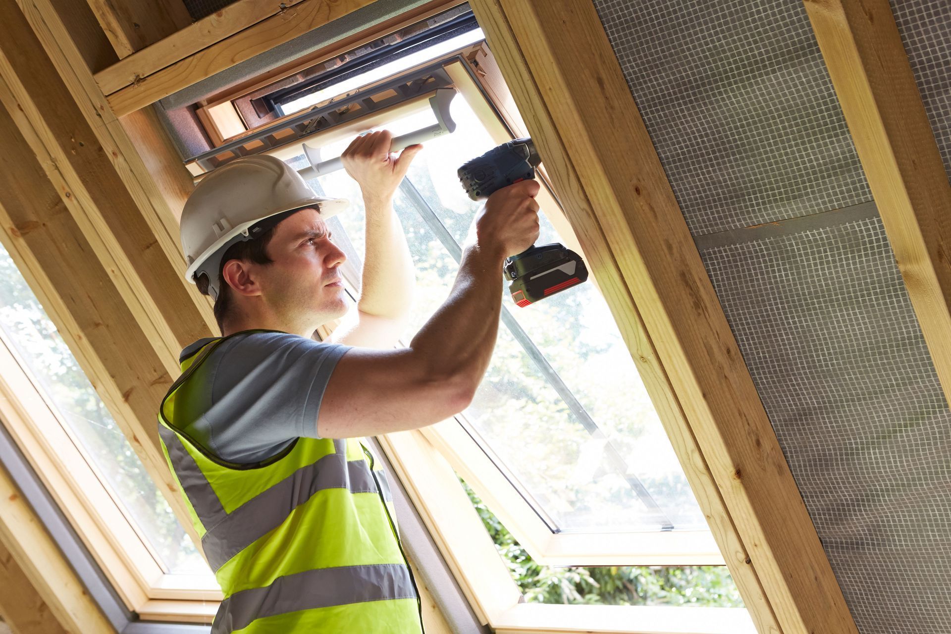 Worker in safety vest and hard hat installing a window frame with a drill in a wooden structure