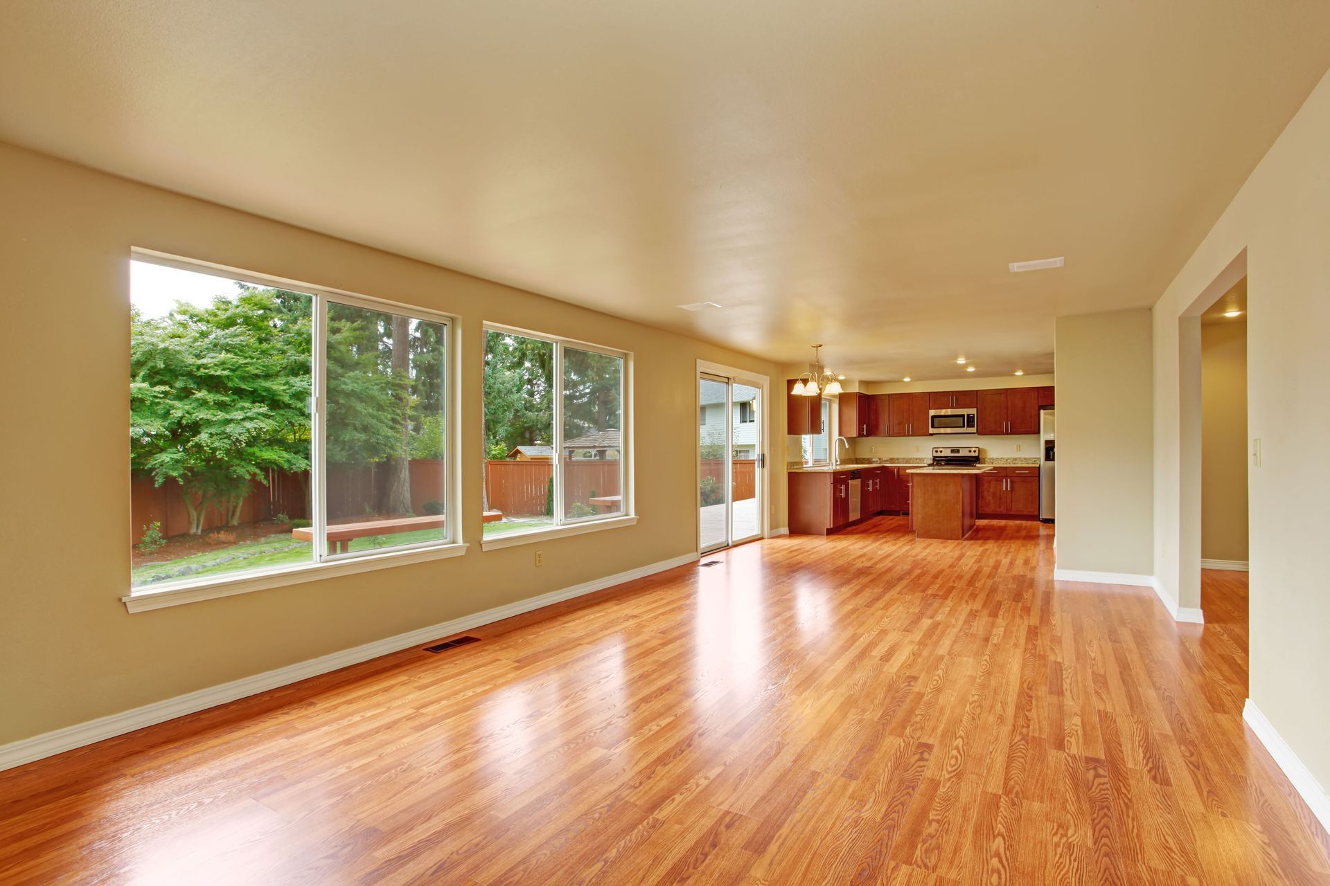Bright empty living room with hardwood floors, large windows, and an open kitchen area
