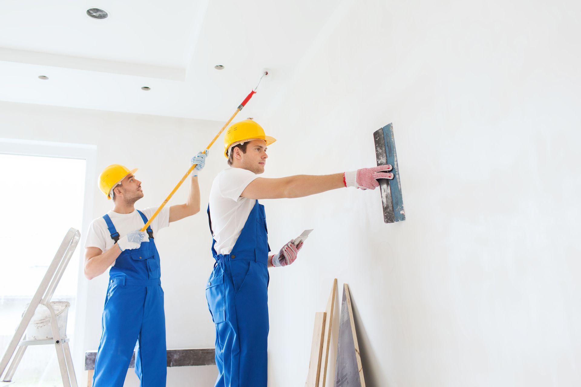 Two workers in blue overalls paint a white wall, one rolling paint while the other uses a tray and extension pole.