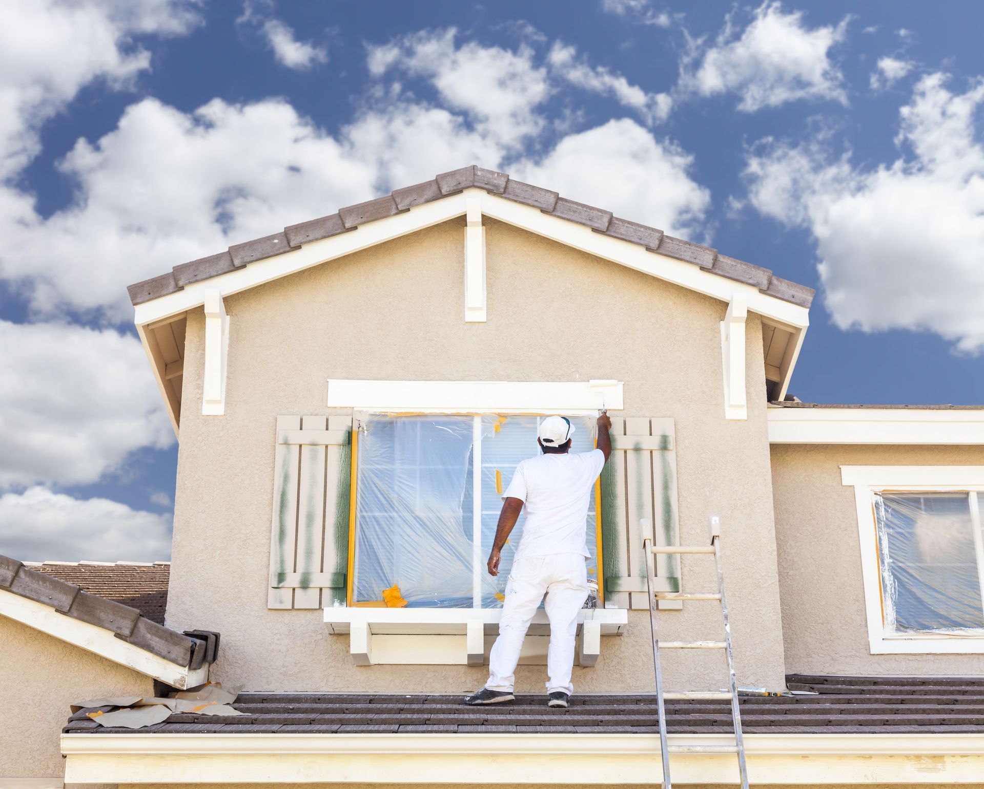 Painter in white workwear painting a house exterior under a cloudy sky, standing on a ladder.