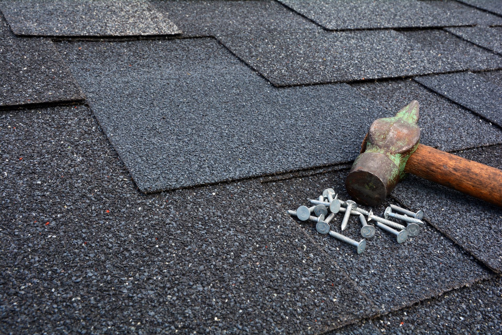 Roof shingles being repaired with a hammer and roofing nails on dark asphalt shingles