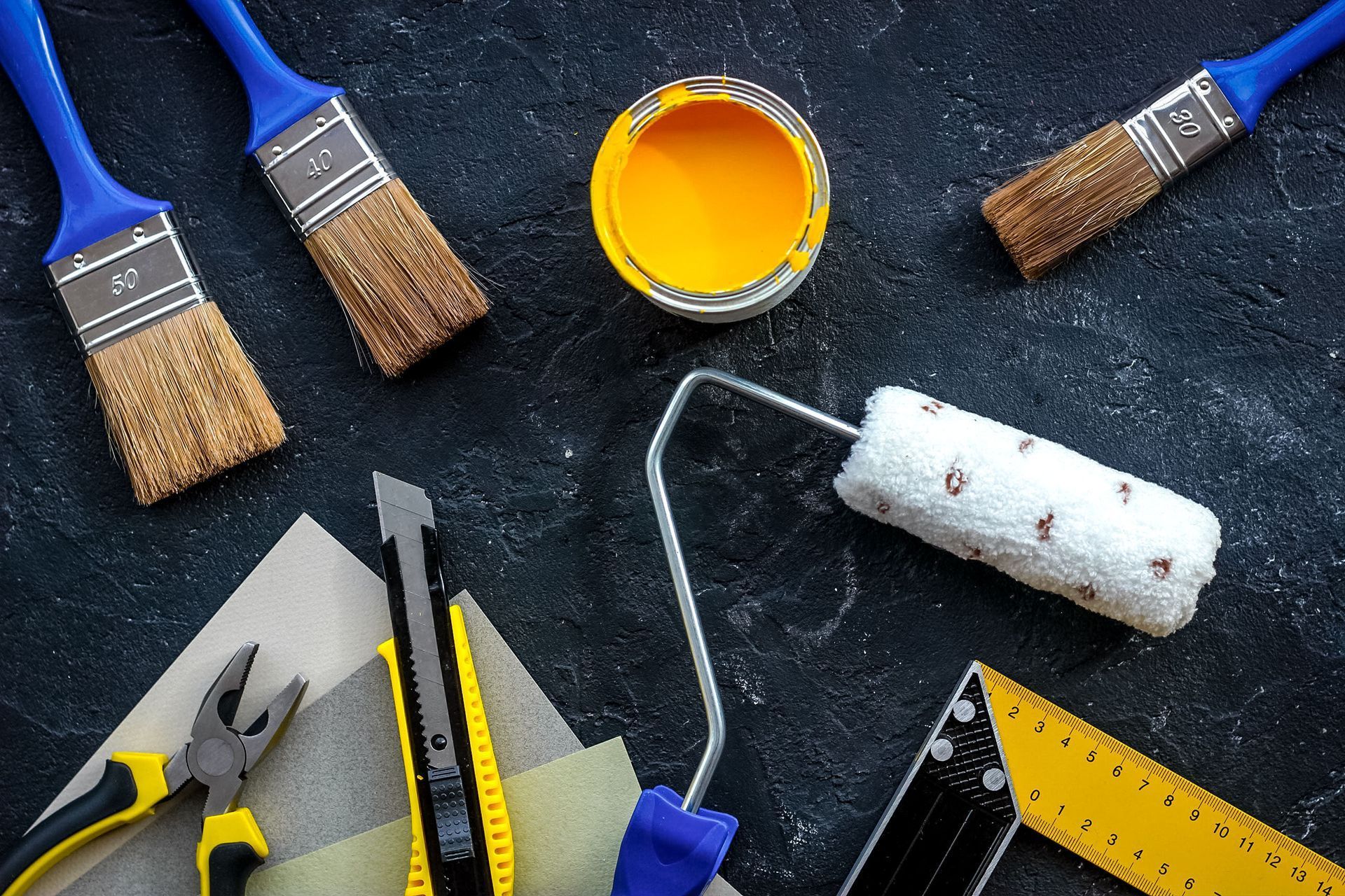 Painting tools and supplies on a dark surface, including brushes, roller, paint can, and tape measure