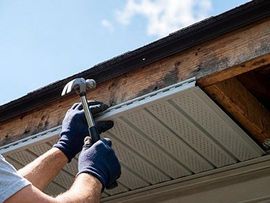 Gloved hands hammering a nail into soffit beneath a house roof eave