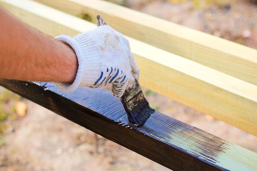 Gloved hand painting a dark wooden board with a brush outdoors