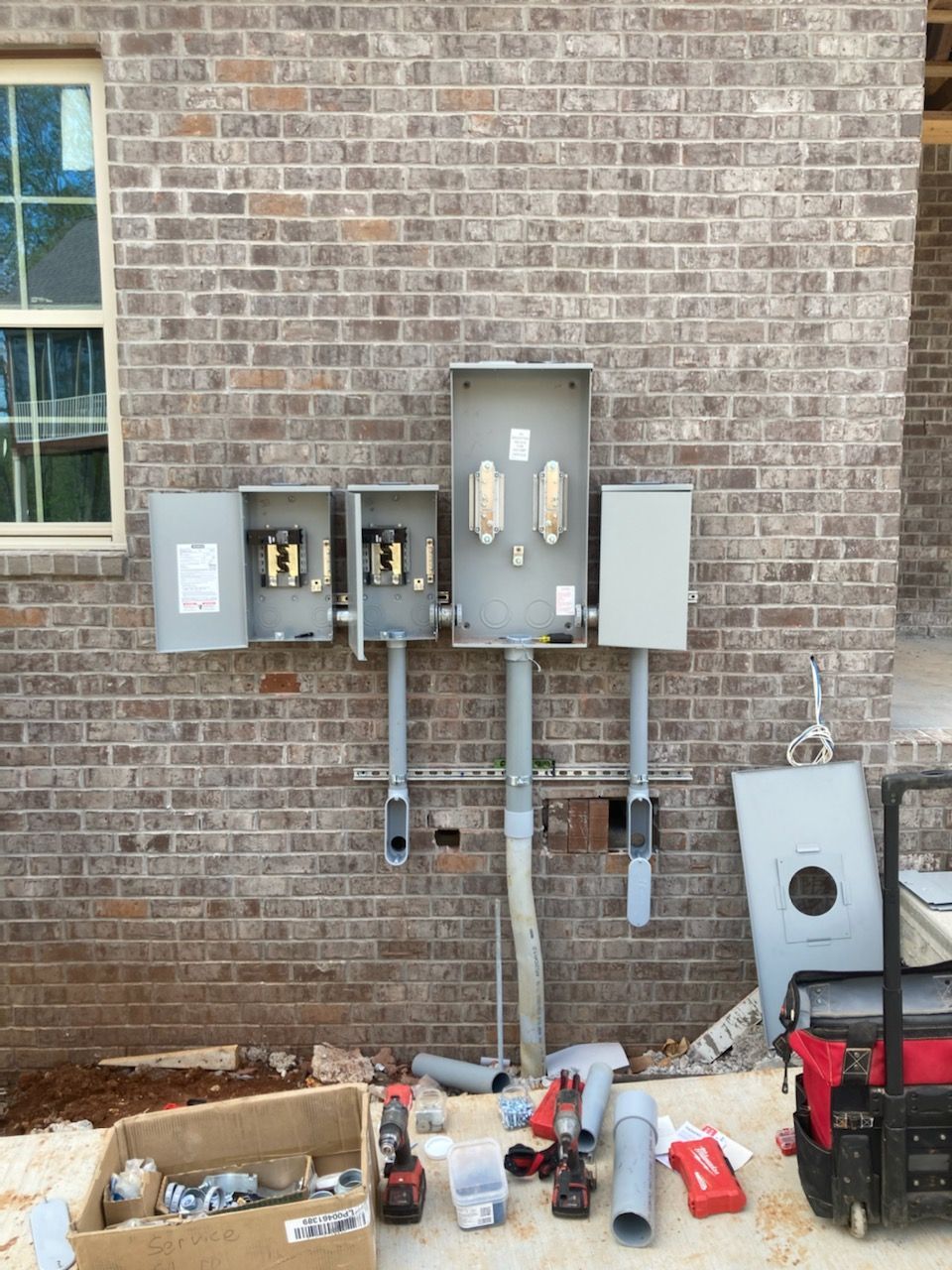 Electrical meters and utility boxes mounted on a brick wall, with tools and supplies on the ground below.