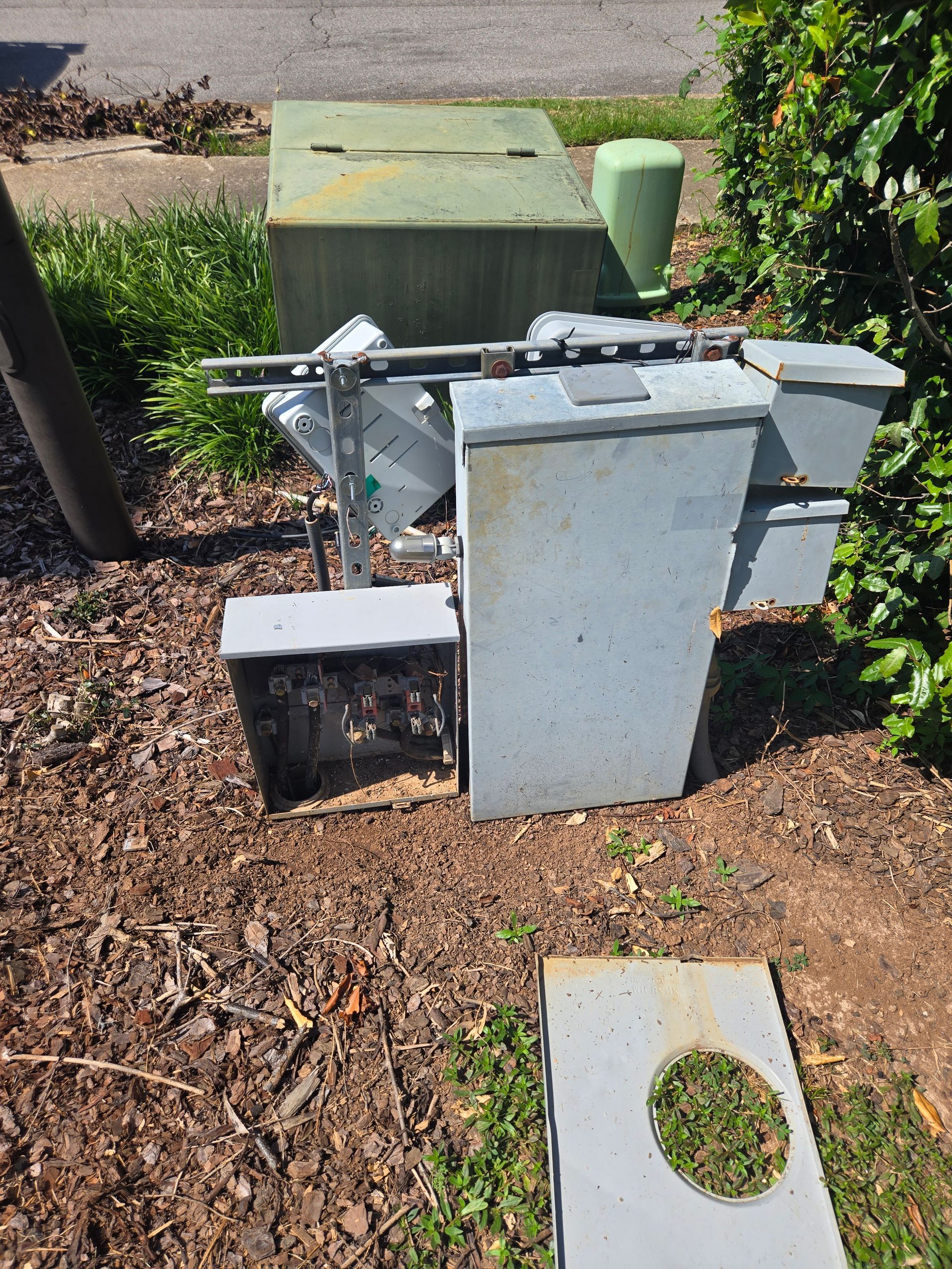 Damaged electrical equipment on the ground next to a curb with vegetation. Gray metal boxes and green top.