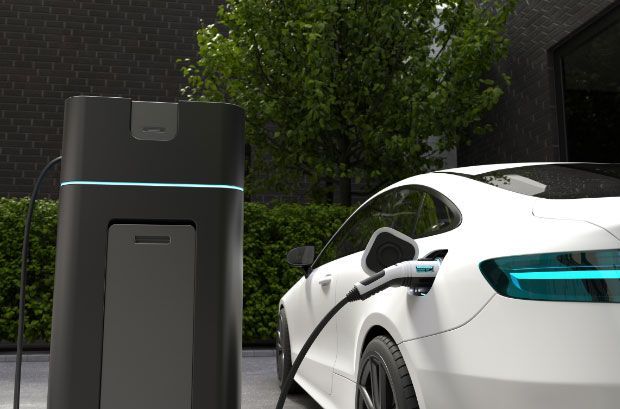 White electric car charging at a black home charging station, next to a green hedge.