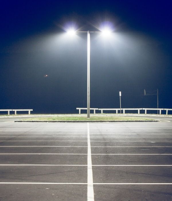 Empty parking lot at night, brightly lit by overhead lamps; benches and a basketball hoop are in the background.