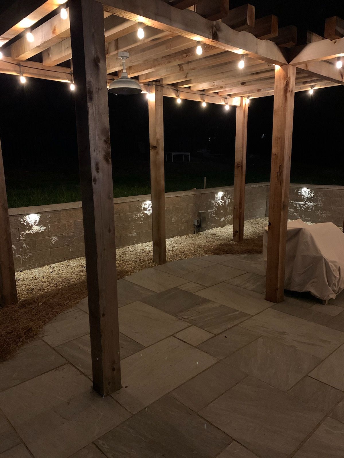 Covered patio with wooden posts, string lights, stone wall, and covered grill at night