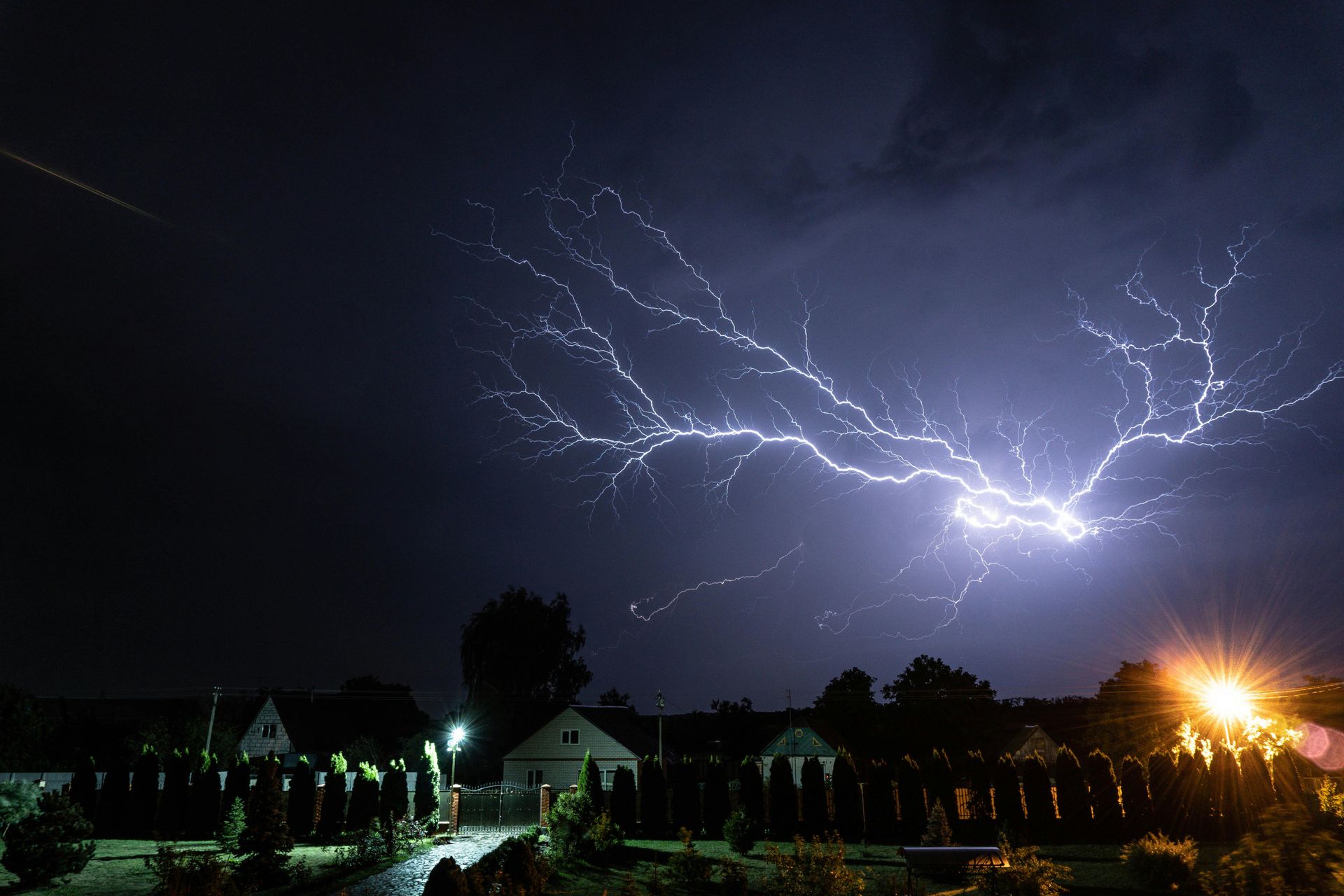 Lightning bolts illuminate a dark night sky over houses and trees with city lights below