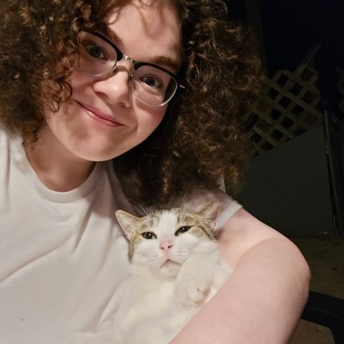 Smiling person with curly hair and glasses holding a white cat indoors at night