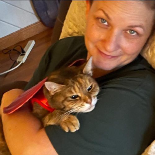 Woman holding a brown tabby cat in a red harness, both looking up at the camera indoors.