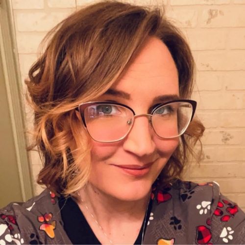 Portrait of a smiling person with glasses and a floral top against a brick wall