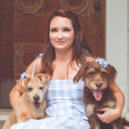 Woman in a blue plaid dress holding two small dogs, one tan and one brown, in front of a wooden door