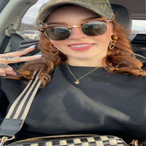 Woman in sunglasses and cap sitting in a car, smiling and posing with a peace sign.