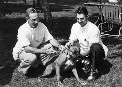 Two people crouch beside a dog on grass in a black-and-white outdoor photo.