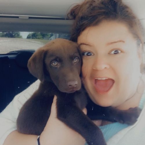 Woman smiling and holding a brown puppy in a car, both looking at the camera.