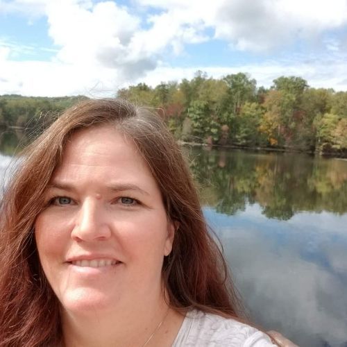 Woman smiling by a calm lake with trees and cloudy sky reflected in the water