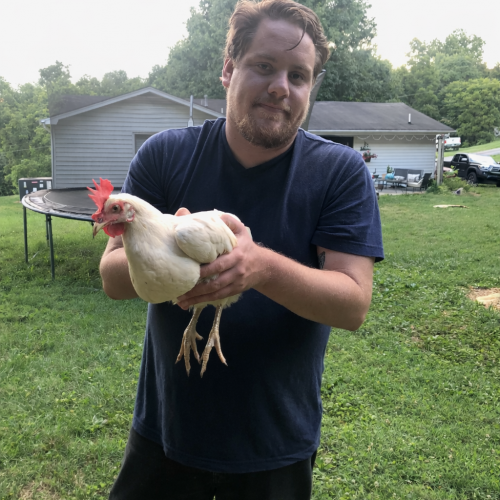 Man holding a white chicken in a grassy backyard with sheds in the background