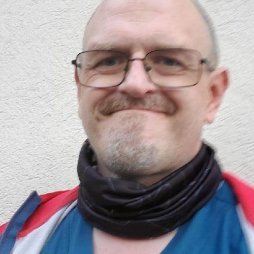 Close-up of a smiling bald man wearing glasses, a black neck gaiter, and a blue shirt with red straps.