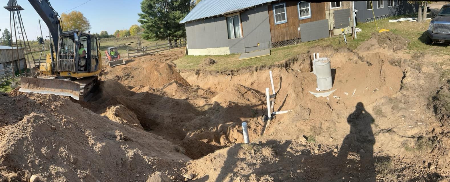 An excavator parked near a house with an excavated trench and exposed sewer pipes in a dirt yard.