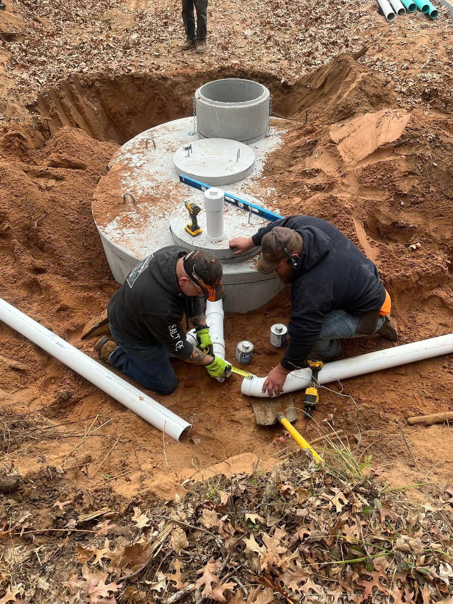 Two people in work gear install white PVC pipes into a concrete septic tank in a dirt excavation site.