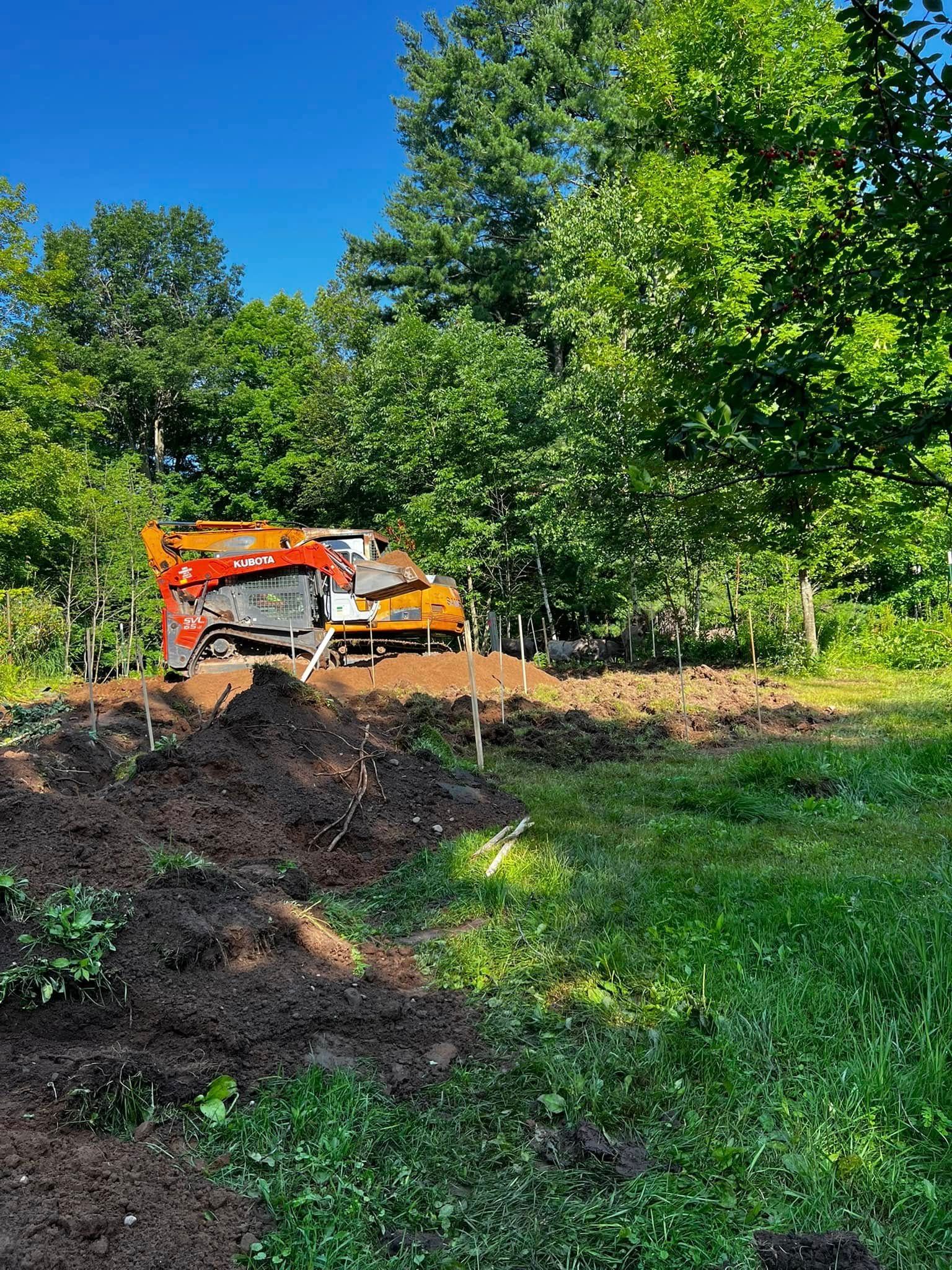 An orange industrial wood chipper sits on a mound of dirt in a sunny, tree-lined clearing.