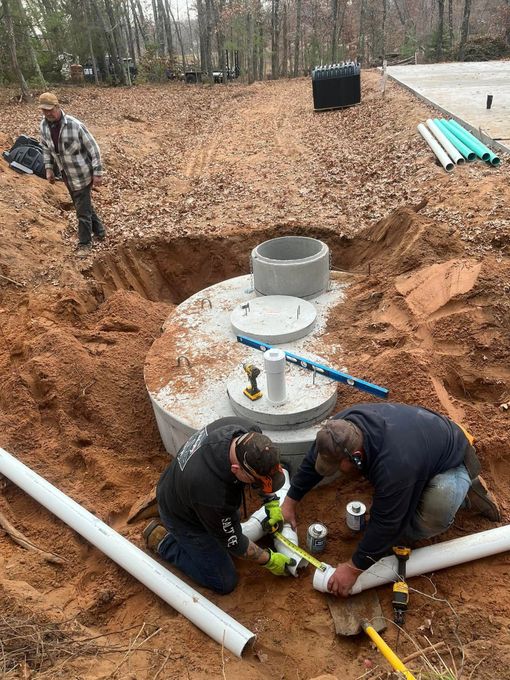 Two workers in a dirt pit installing plumbing pipes into a circular concrete septic tank on a construction site.