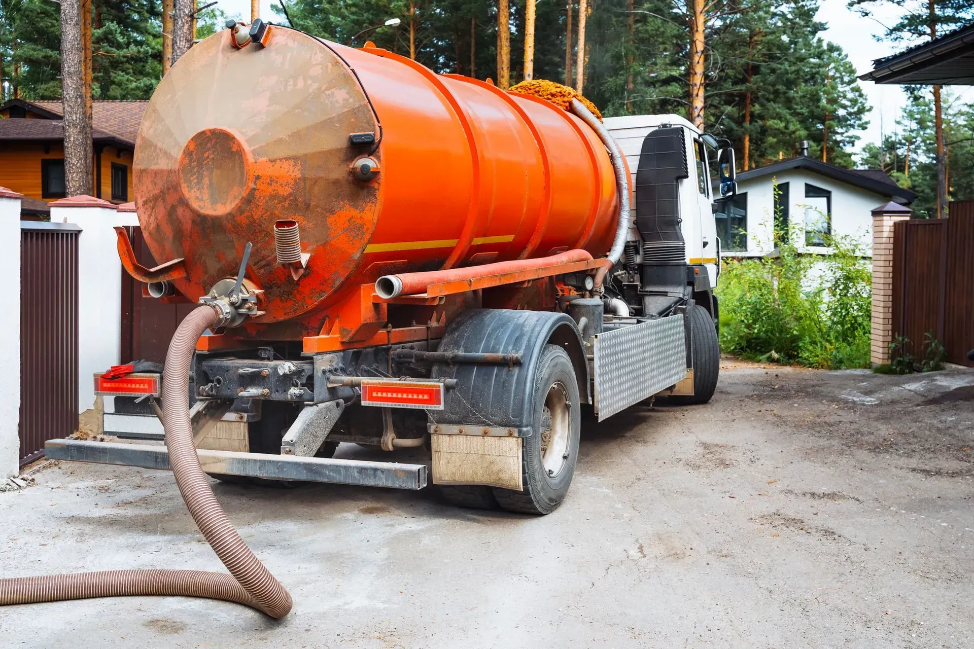 A bright orange vacuum truck parked on a paved driveway in front of houses and trees, with a large hose extended.