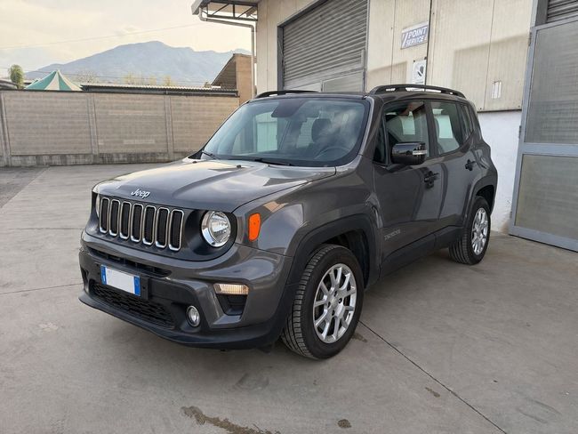 Una Jeep Renegade grigio scuro è parcheggiata fuori da un edificio su un piazzale di cemento, con le montagne sullo sfondo.