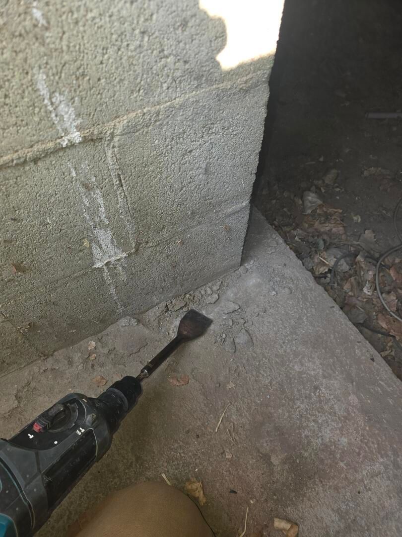 Black handheld tool pointed at a concrete wall corner on a floor in a dim, unfinished space