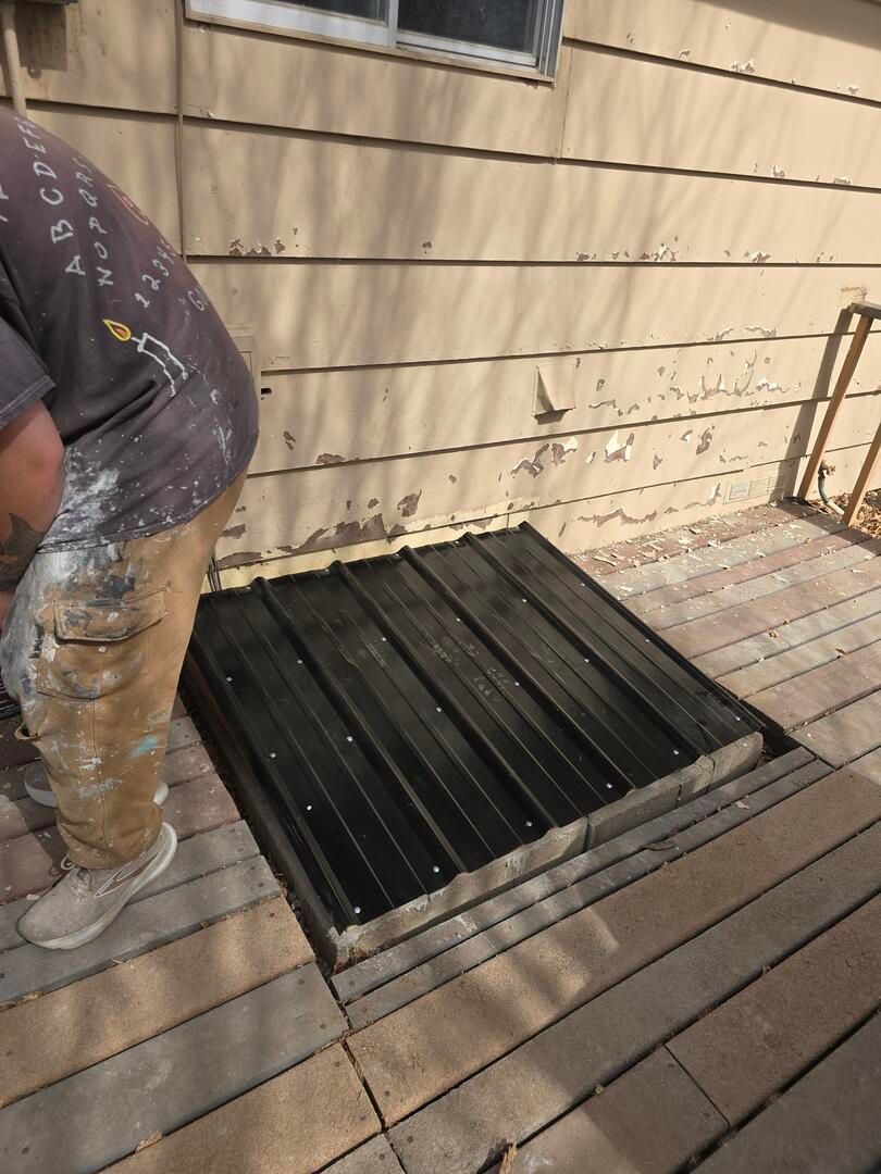 Worker installing a black metal vent on a wooden deck beside a house wall.