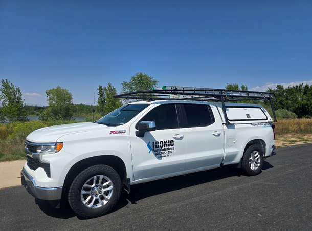 White work pickup truck with Iconic Pest Solutions labeled on the side utility canopy and ladder rack parked on roadside under a blue sky in colorado