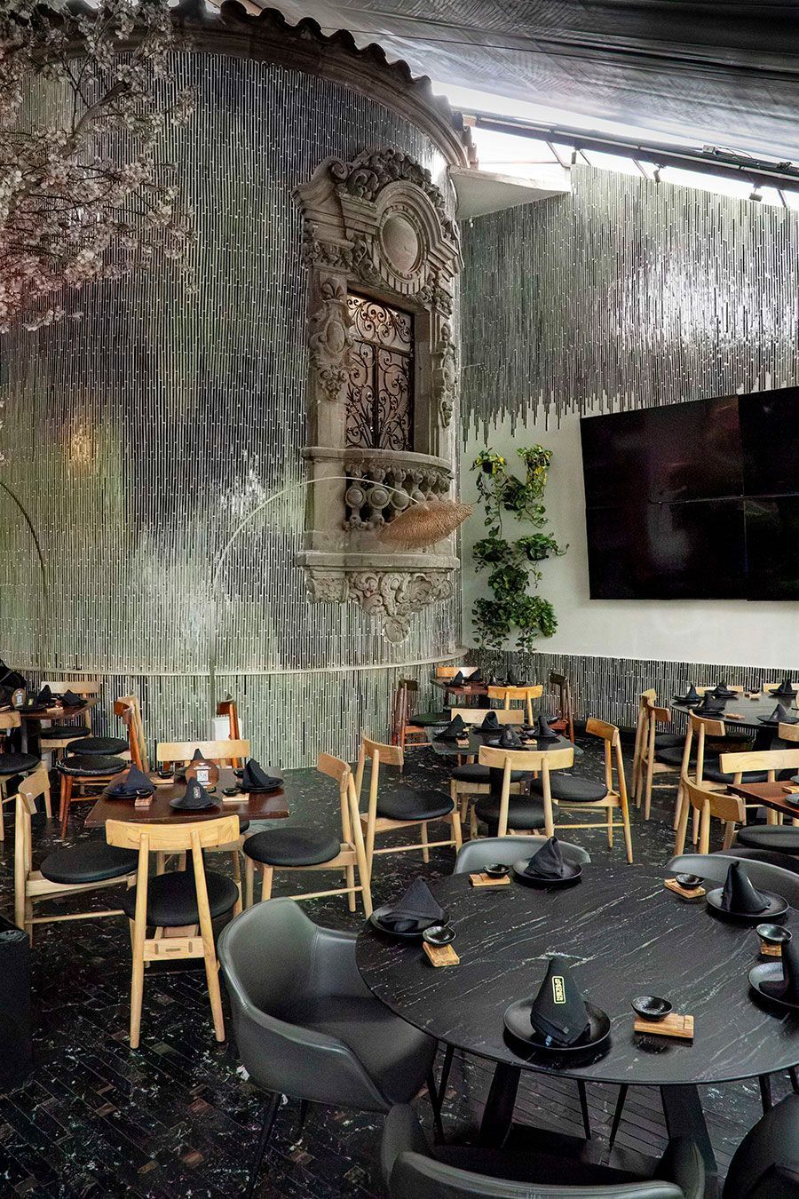 Interior de restaurante moderno con mesas de madera, sillas negras y altos árboles de interior bajo un techo alto.