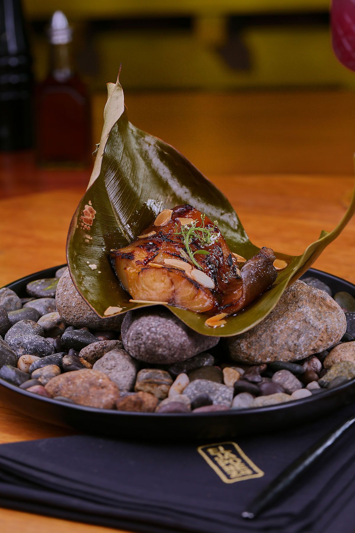 Pescado a la parrilla envuelto en una hoja verde, servido sobre una cama de piedras en un plato negro.