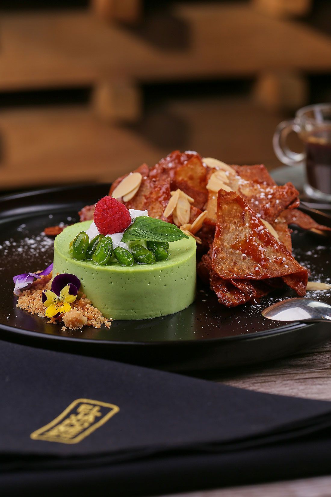Plato de postre con mousse verde, frutos rojos y guarnición de caramelo crujiente sobre una mesa oscura de restaurante.