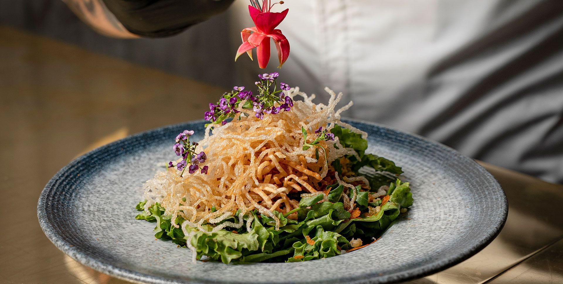 Ensalada gourmet con fideos crujientes, flores moradas y verduras en un plato con borde azul.