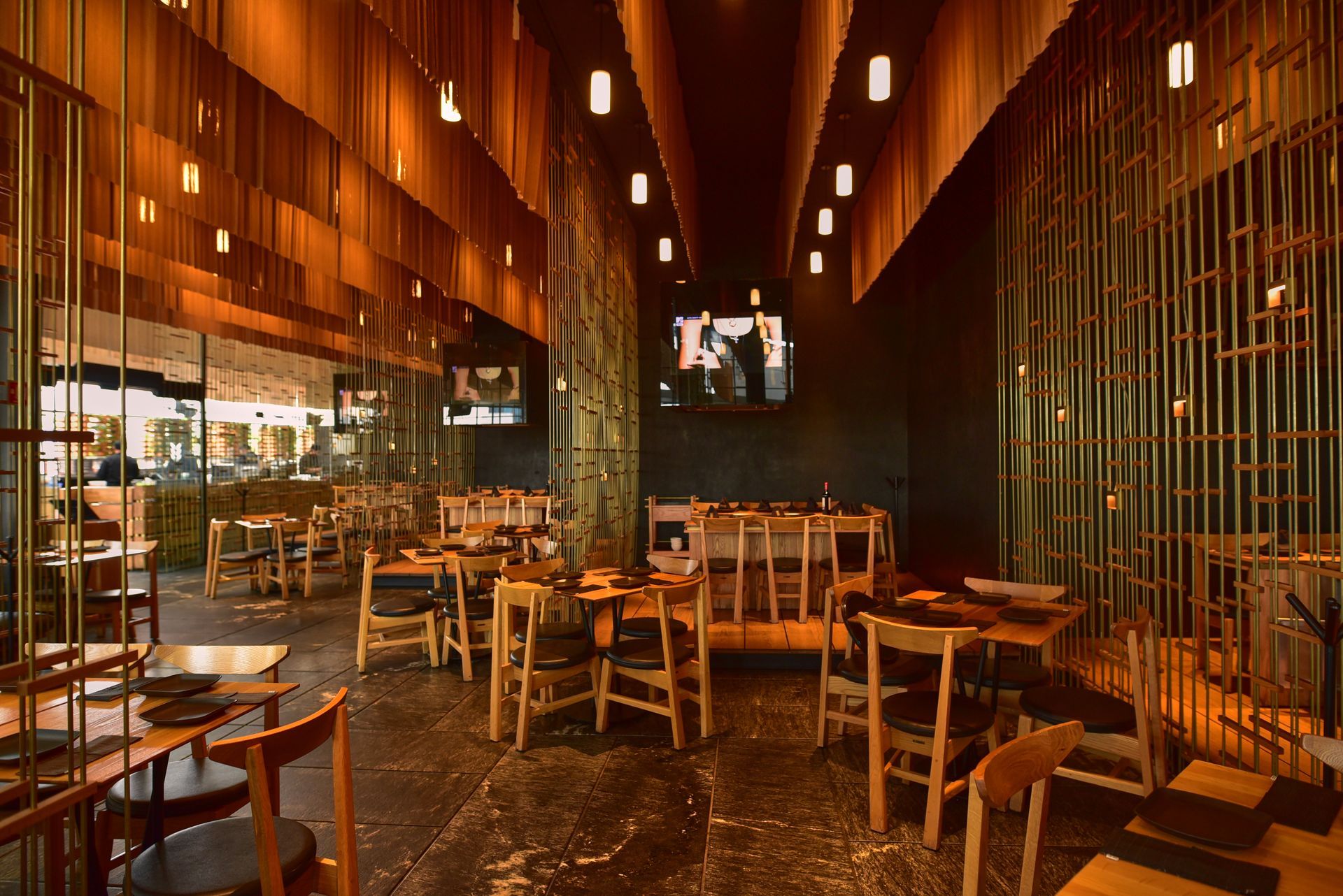 Interior de restaurante moderno con iluminación cálida, sillas y mesas de madera y lámparas colgantes.