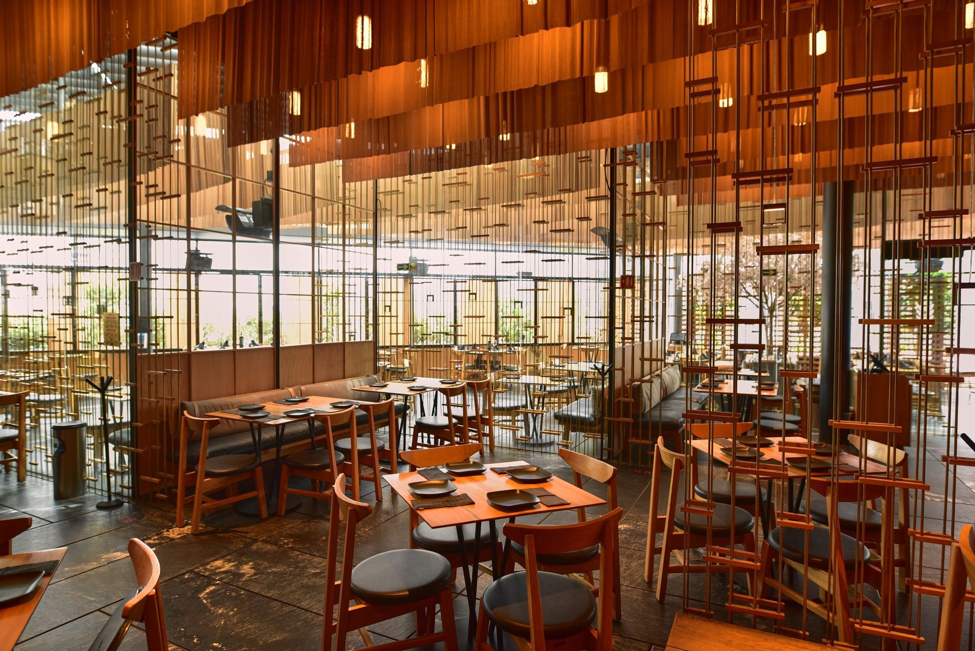 Interior de restaurante moderno con mesas y taburetes de madera, e iluminación cálida mediante lámparas colgantes junto a grandes ventanales.